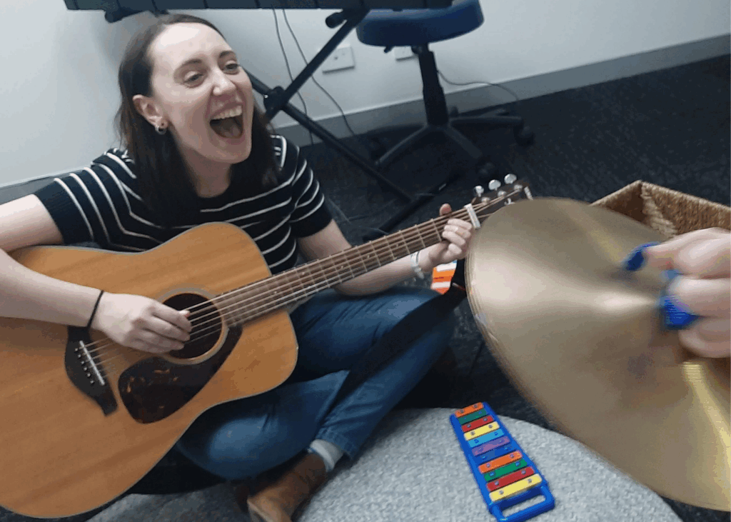 A woman sits on the floor laughing whilst playing an acoustic guitar. In the foreground, someone holds cymbals, and a colourful xylophone is visible—a lively scene you might find at an OT Gold Coast or Gold Coast speech therapist session.