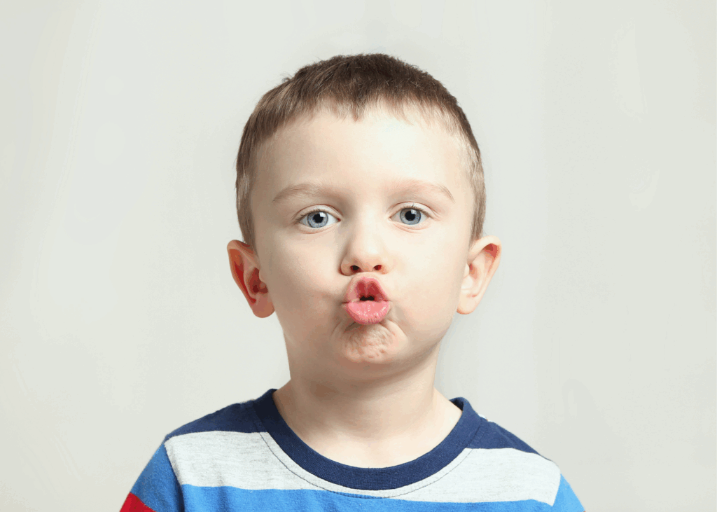 A young boy with light skin and short brown hair makes a fish face by puckering his lips. He wears a striped shirt of blue, white, and gray, perhaps practicing speech therapy for kids, standing against a plain light background.