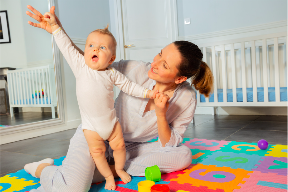 A smiling woman sits on a colourful foam play mat, holding up a happy baby who is reaching upwards. Toys and a cot are visible in the bright room, ideal for OT Gold Coast feeding therapists to support child development and play.