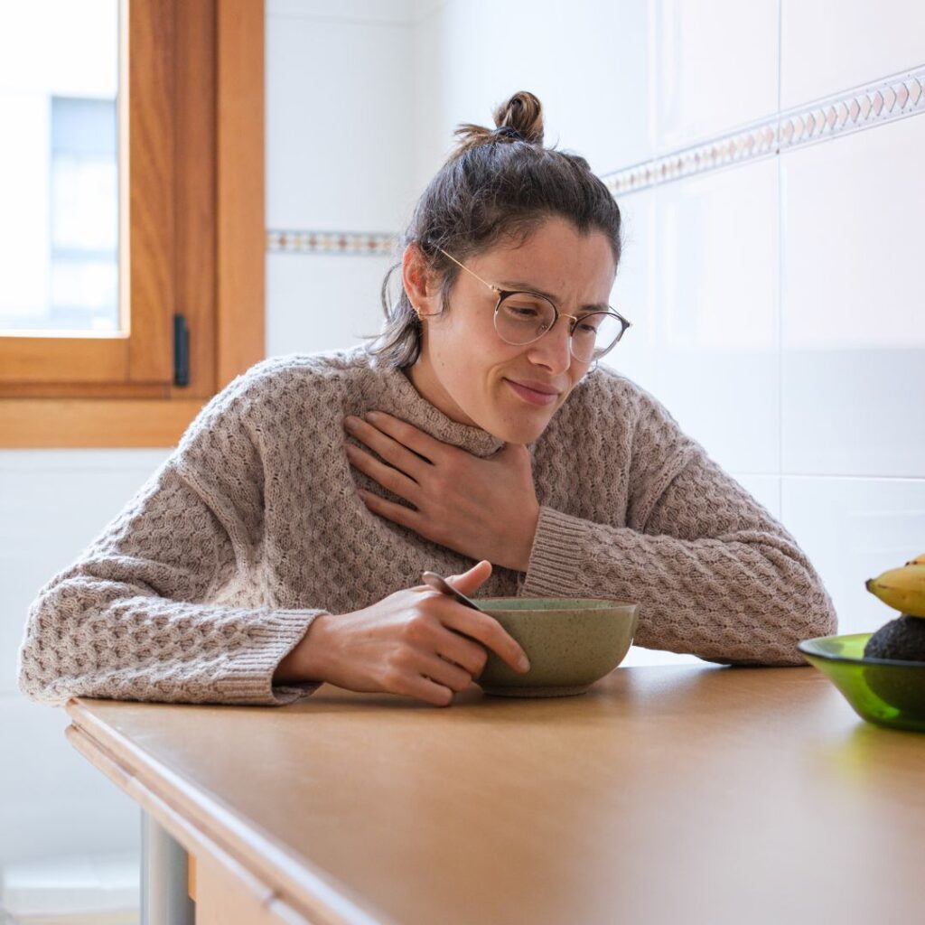 A woman wearing glasses and a knitted jumper sits at a kitchen table with a bowl in front of her, holding her hand to her throat and appearing uncomfortable—possibly experiencing throat pain or needing a Gold Coast speech therapist.