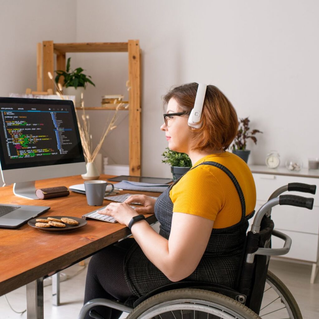 Lady in wheelchair with headphones on sitting in wheelchair at her computer desk, looking at changes to her NDIS plan's funding period.