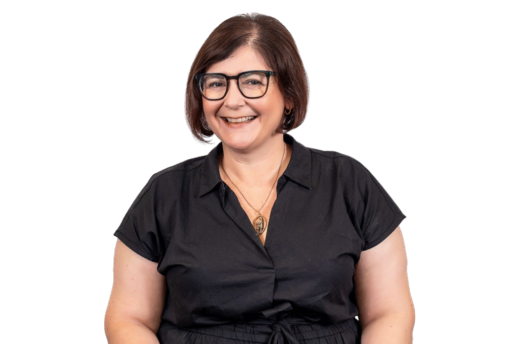 A woman with short brown hair and glasses smiles while wearing a black short-sleeve shirt and gold necklace, posed against a plain white background—perfect to represent music therapy courses or a music therapist Gold Coast professional.