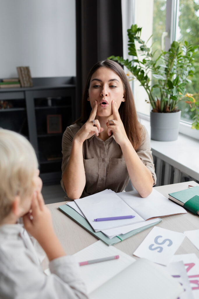 A woman sits at a desk with open notebooks and letter cards, demonstrating a mouth movement by touching her cheeks with her fingers, while a child watches and imitates her during speech therapy for kids.