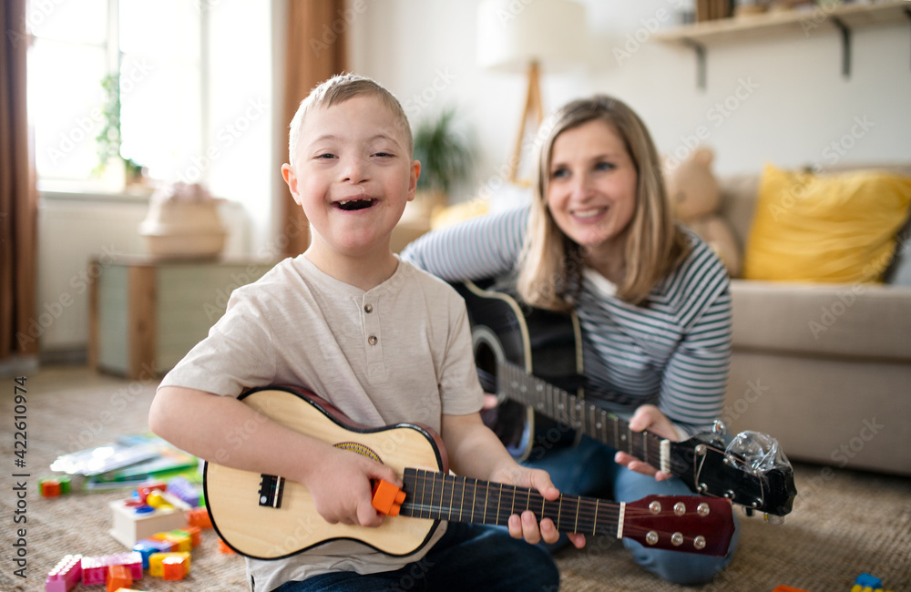A smiling boy with Down syndrome sits on the floor playing a small guitar, while his music therapist from Gold Coast smiles behind him, holding a guitar. Colorful toys are scattered around them in their cozy living room.