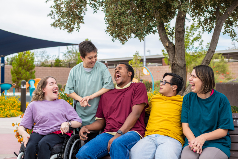 A diverse group of five friends, including one person in a wheelchair, sit on a bench outdoors, enjoying laughter and smiles together under a tree—reflecting the inclusive spirit of music therapy Gold Coast.