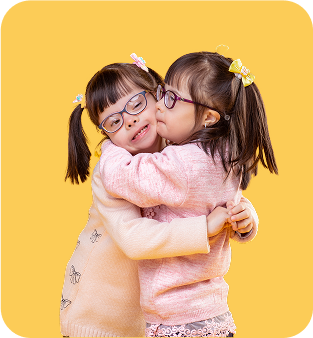 Two young girls with glasses and pigtails, wearing pastel sweaters, hug each other closely and smile in front of a solid yellow background—radiating the warmth of friendship found in music therapy gold coast sessions.