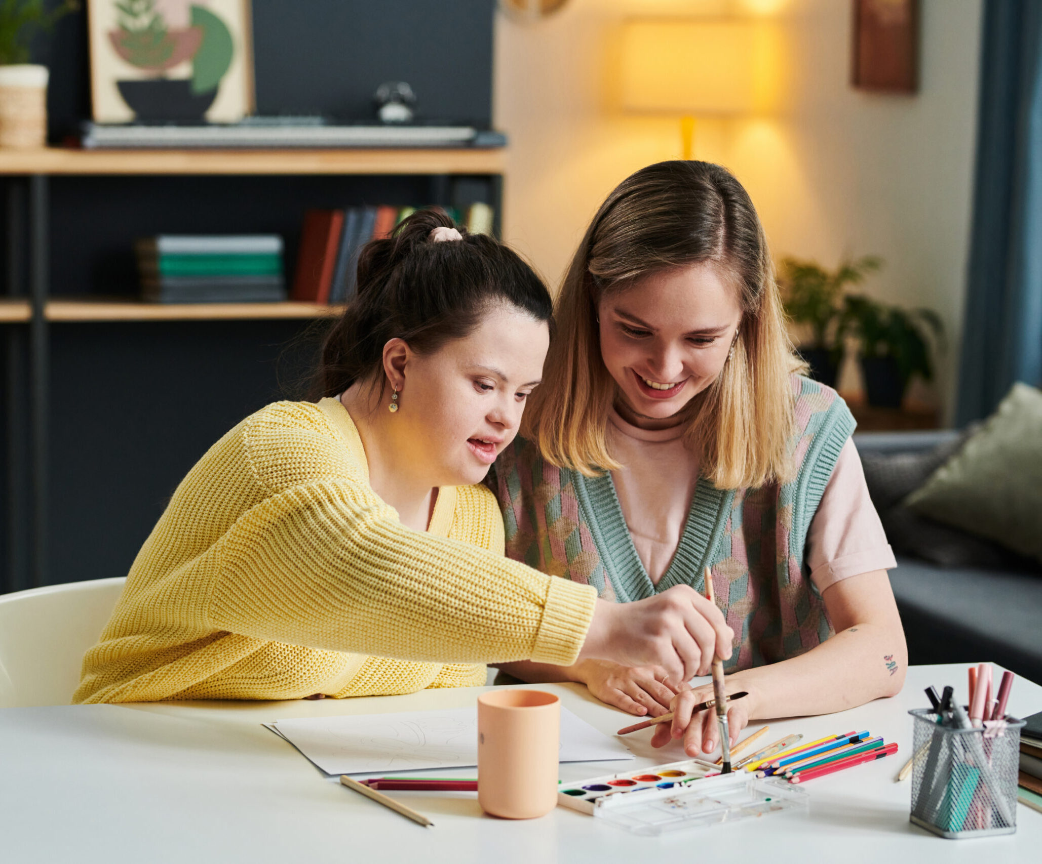 Two young women sit at a table, smiling and drawing together with colored pencils. Art supplies, paper, and a mug are on the table in a cozy, well-lit room—perfect for those interested in music therapy courses or creative therapies.