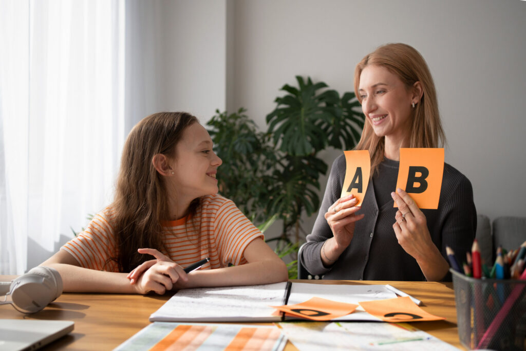 A woman and a girl sit at a table with books and papers. The woman, a music therapist Gold Coast, smiles as she holds up two cards labeled A and B, while the girl looks at her and smiles during their session.