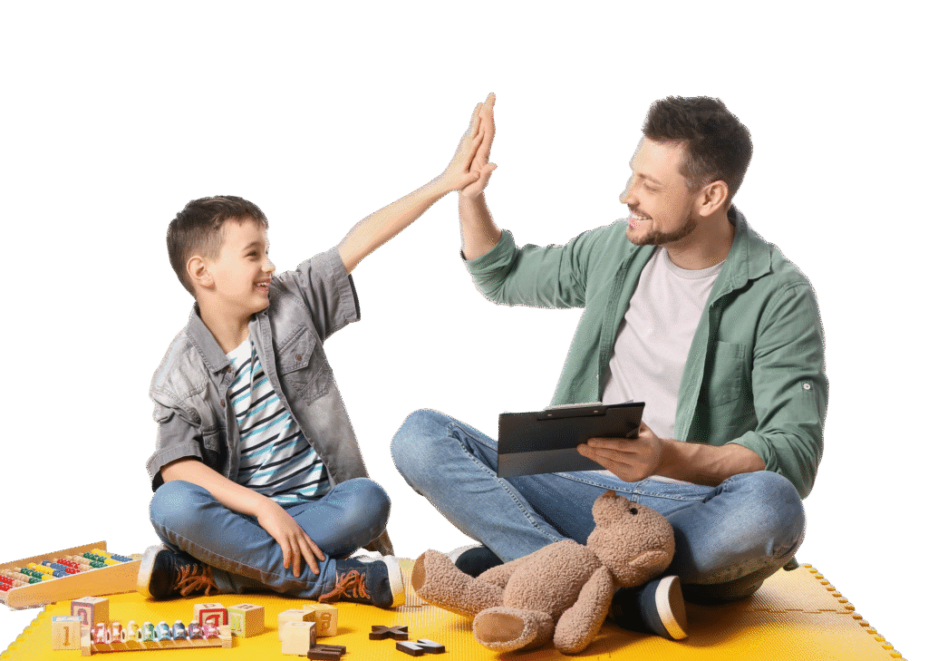 A man and a boy sitting on a yellow mat give each other a high-five. The man holds a tablet, and toys and a teddy bear are in front of them. Both are smiling, enjoying their time together—perhaps during speech therapy for kids.