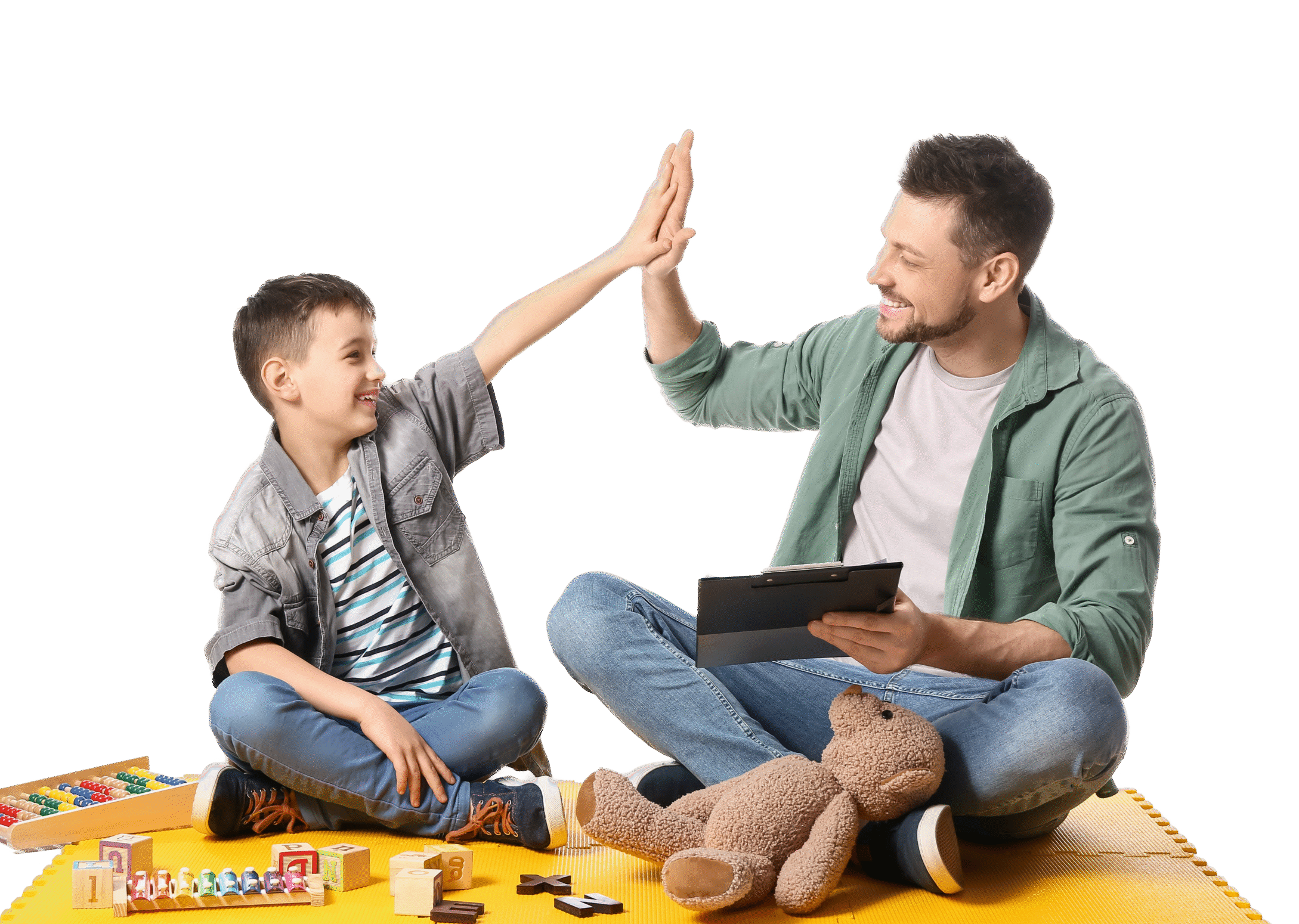 A man and a boy sitting on a yellow mat give each other a high-five. The man holds a tablet, and toys and a teddy bear are in front of them. Both are smiling, enjoying their time together—perhaps during speech therapy for kids.