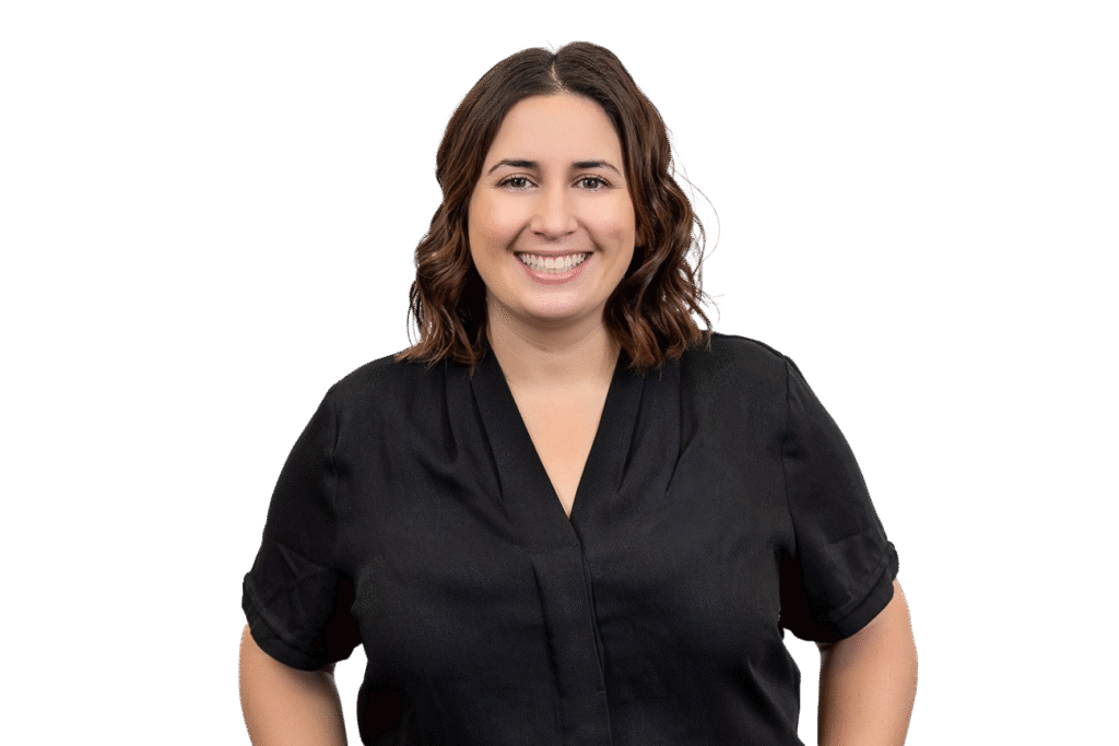 A woman with wavy brown hair, wearing a black short-sleeve blouse, smiles while standing against a plain white background—a welcoming presence for anyone interested in music therapy Gold Coast services.