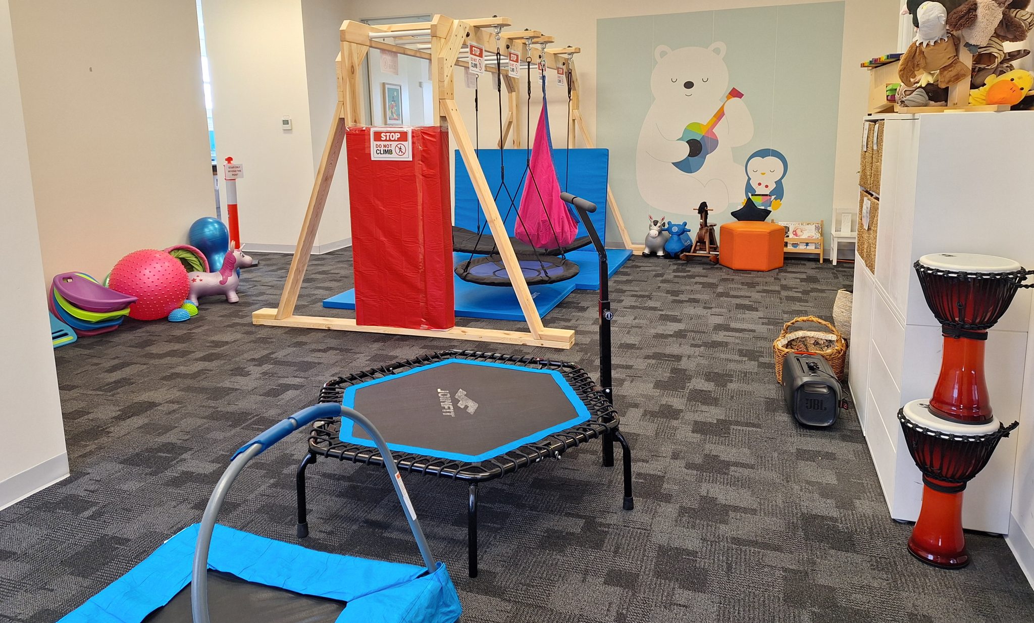 A sensory playroom with swings, a mini trampoline, exercise balls, drums for music therapy gold coast sessions, a climbing frame, and colorful toys. Animal paintings adorn the walls, while shelves hold stuffed animals and sensory equipment.