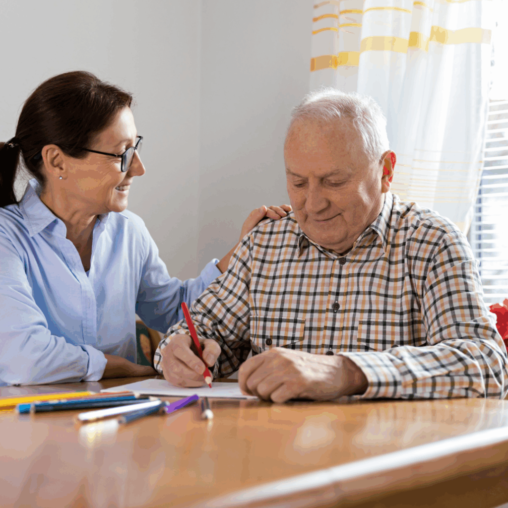 A woman sitting with an older man supporting him to write on paper.