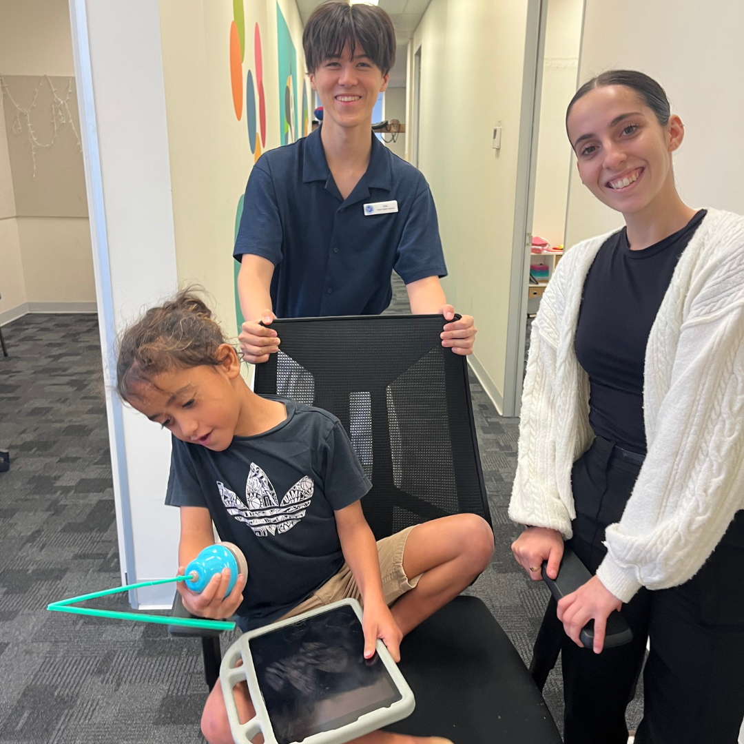 Young boy with a communication device in a chair with a supporting man and woman standing with him.