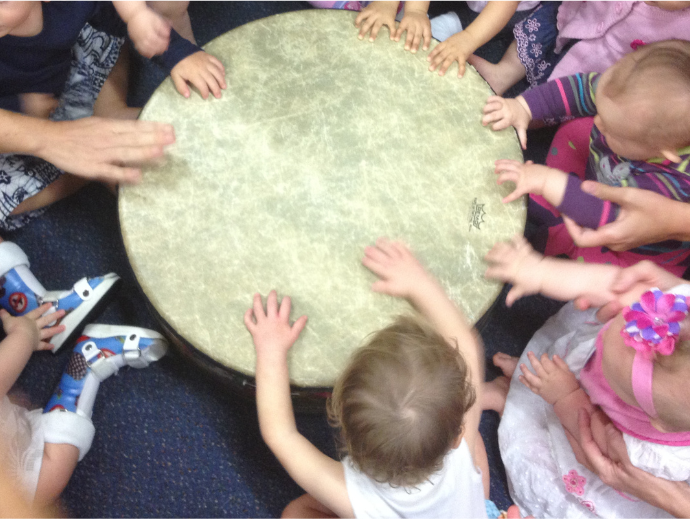 Several young children sit in a circle, reaching out and placing their hands on a large drum, enjoying a playful moment during music therapy gold coast sessions.