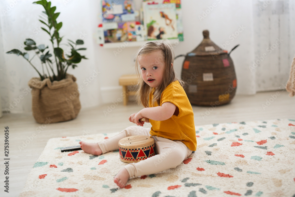 A young girl with Down's syndrome sits on a patterned rug in a bright room, playing a decorative hand drum during music therapy NDIS. She wears a yellow shirt and light trousers, with plants and baskets visible in the background.