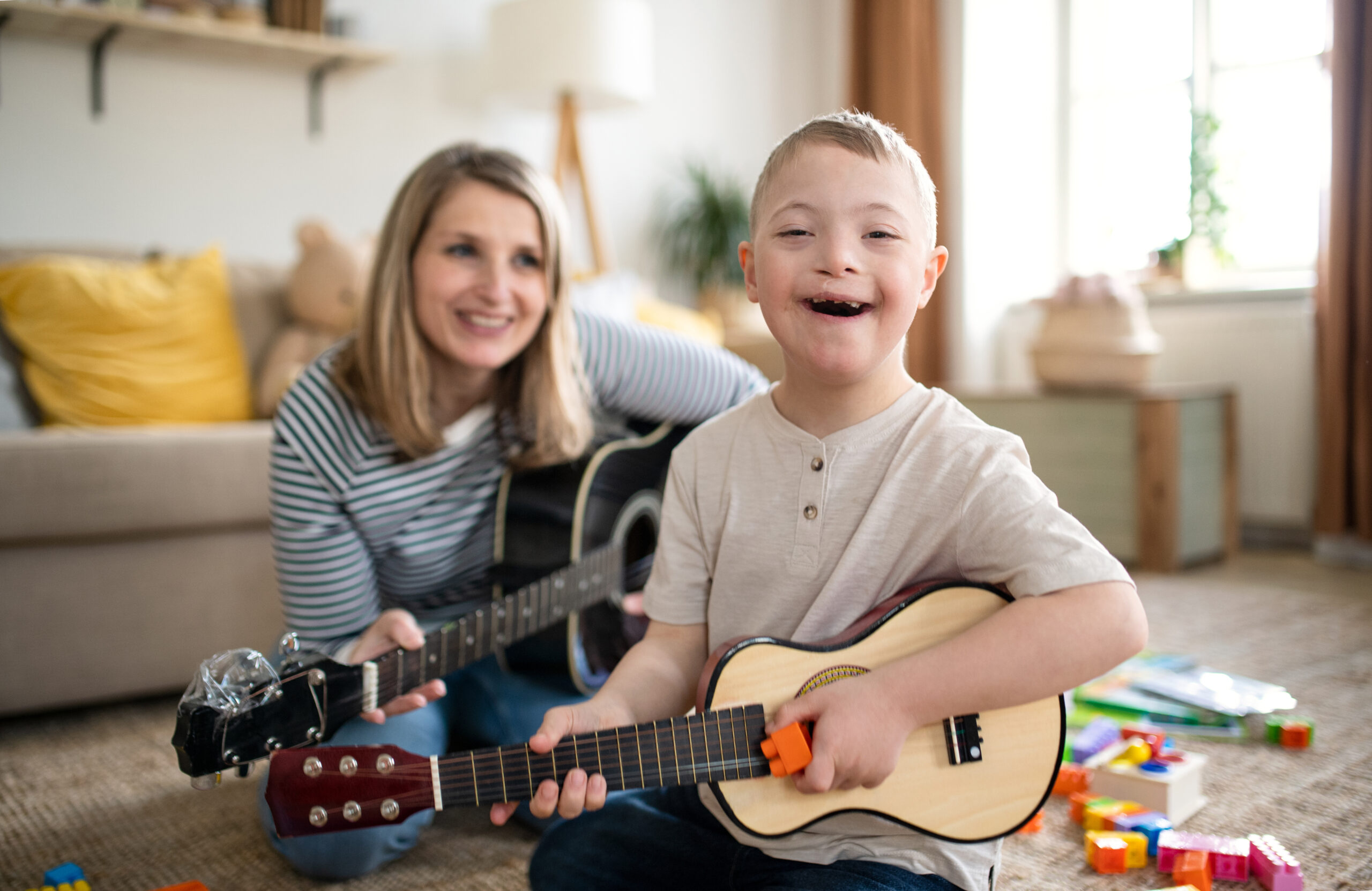 A smiling young boy with Down’s syndrome plays a small guitar, while a woman beside him, also smiling, holds a guitar. Surrounded by colourful blocks, they enjoy music therapy NDIS together in their cosy living room.