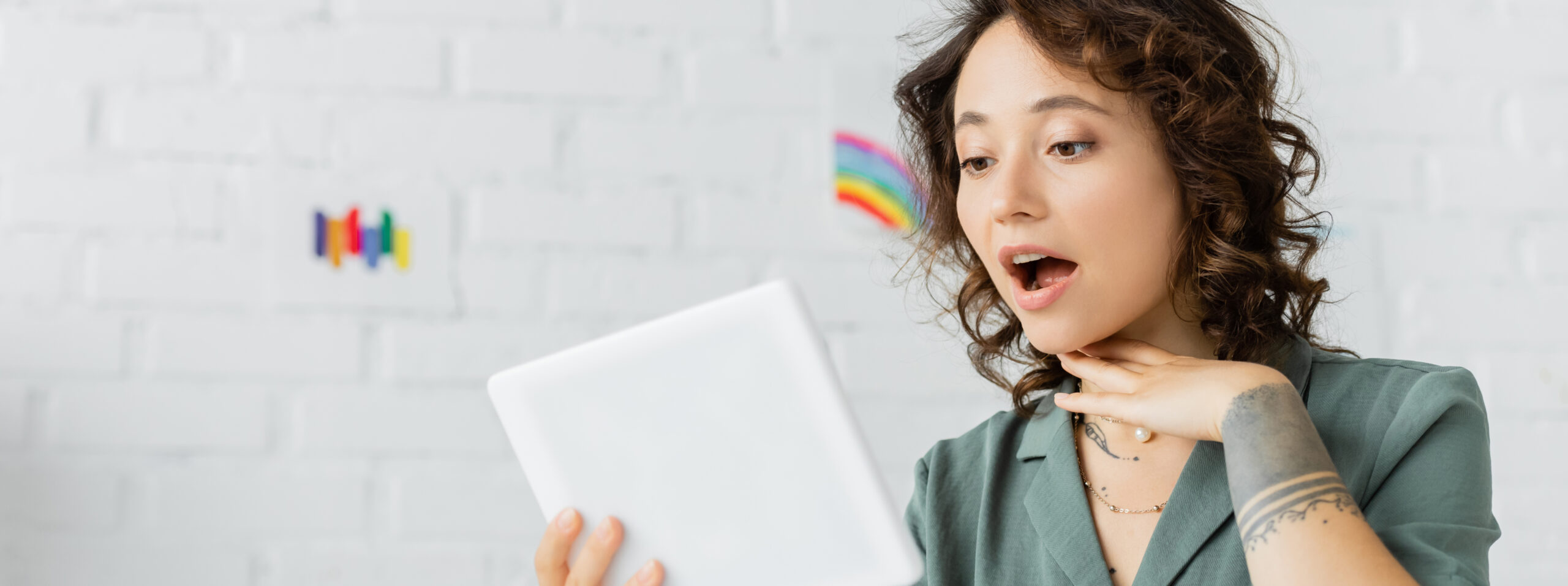A young woman with curly hair looks surprised while holding a tablet. She rests her hand on her chin and sits in front of a white brick wall, possibly awaiting advice from a Gold Coast speech therapist, with a small rainbow drawing in the background.