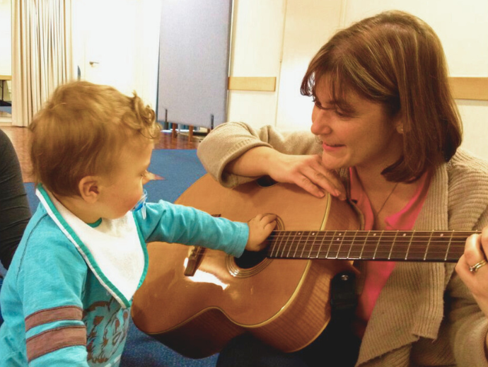 A baby reading out to strum a guitar held by a woman.