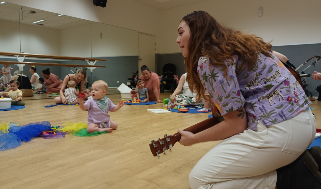 A woman, possibly a music therapist, plays guitar on a wooden floor in a bright room on the Gold Coast, leading babies in playful music therapy with colorful scarves and toys during a lively session with adults.