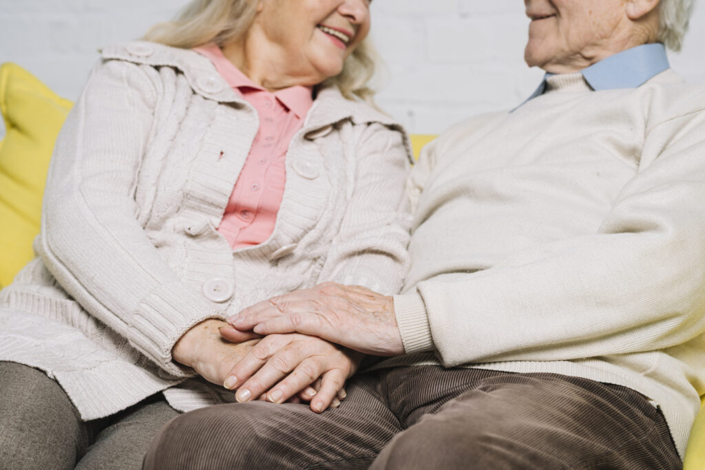 Close-up of an elderly couple sitting together on a yellow couch, smiling and holding hands, wearing cozy sweaters and casual clothes, showing warmth and affection—perfect for promoting music therapy gold coast services.