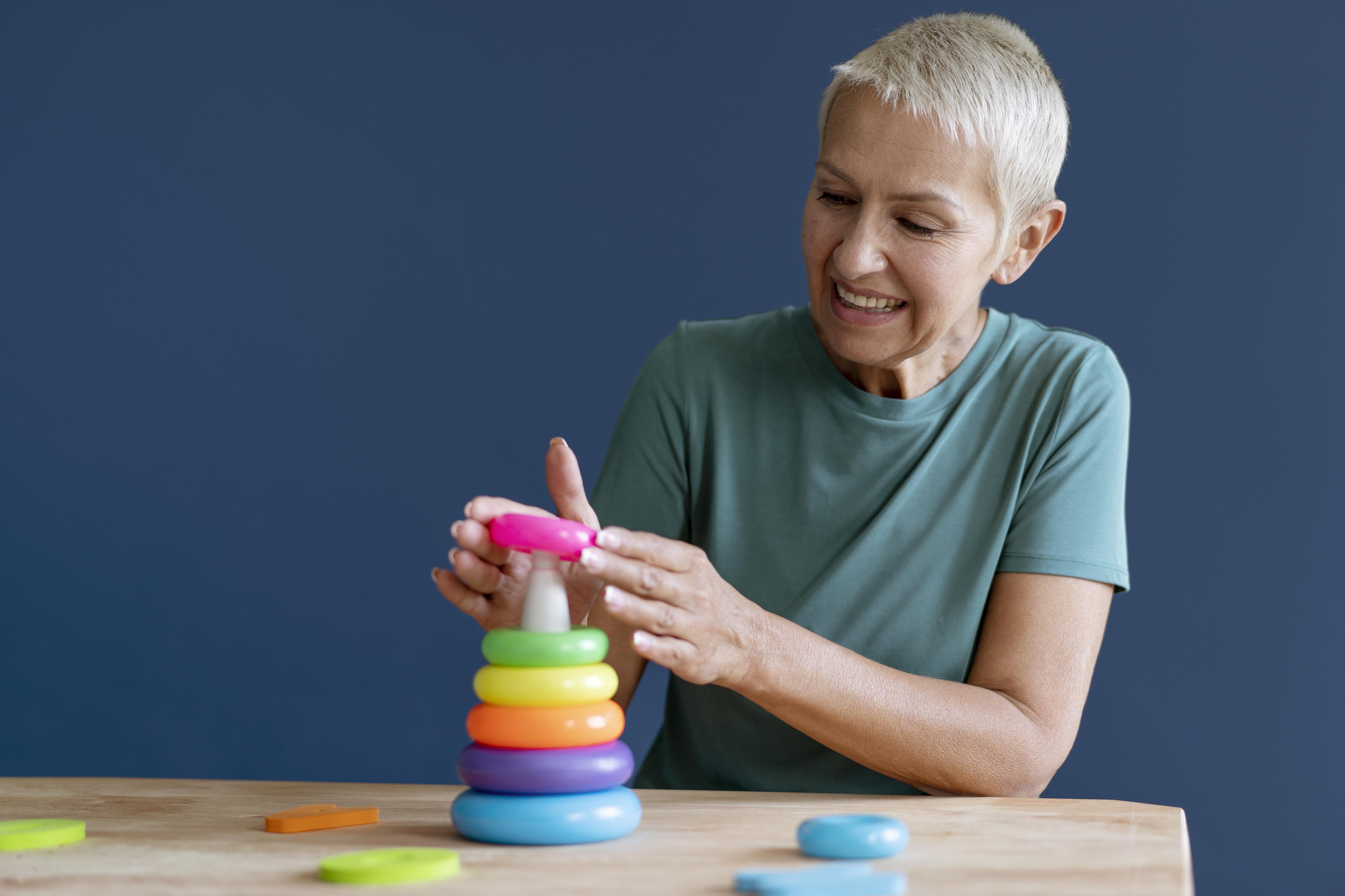 A smiling older woman with short grey hair stacks colourful plastic rings on a wooden table—an activity often used by feeding therapists—against a dark blue background.
