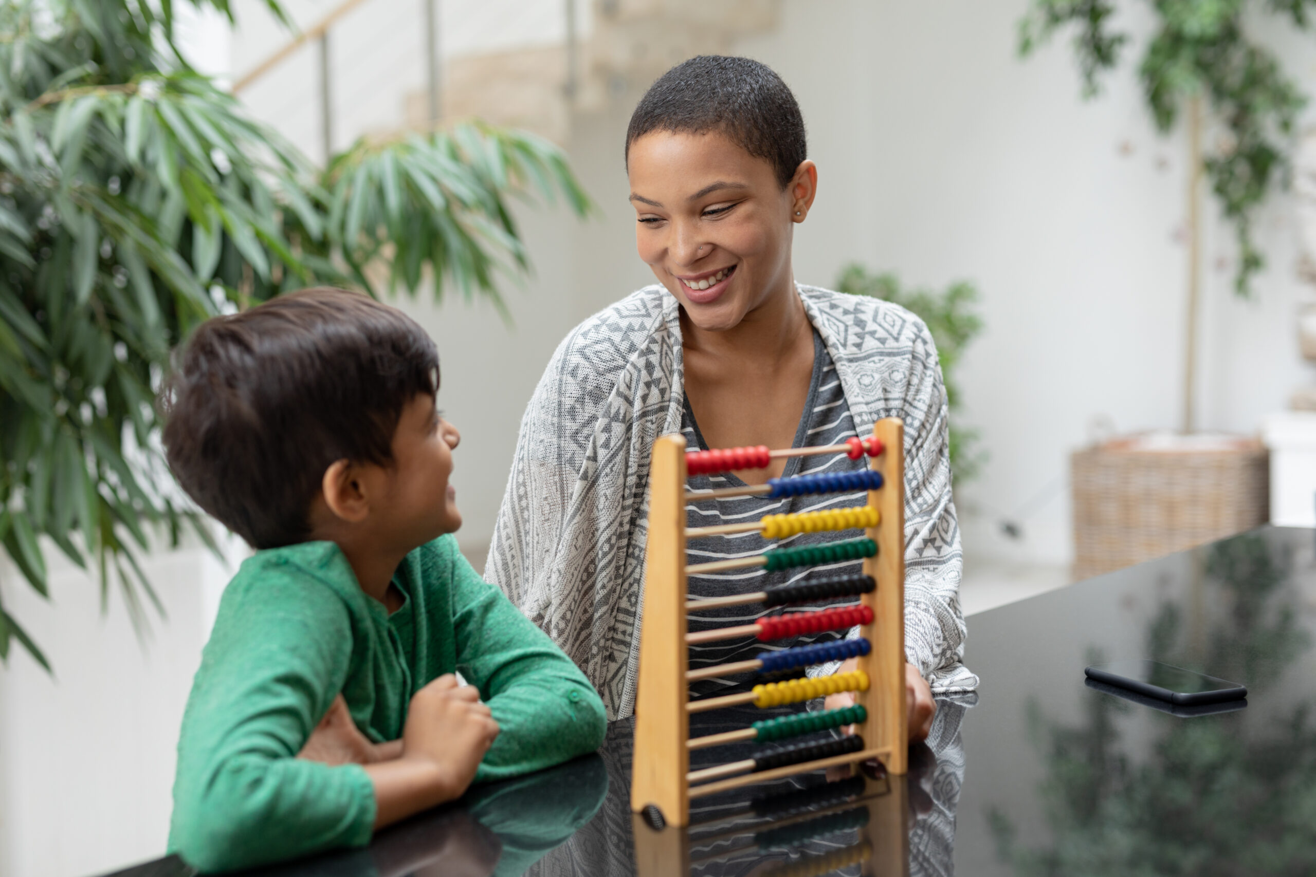 A woman and a young boy sit at a table smiling at each other, with a colourful abacus between them. Lush green plants are in the background, suggesting the welcoming space of feeding therapists or OT Gold Coast specialists.