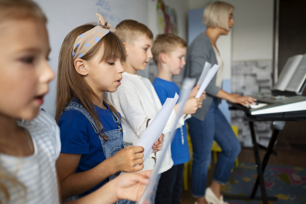 A group of children stand in a row holding sheets of paper and singing, whilst an adult, possibly an OT Gold Coast specialist, plays a keyboard in the background. The setting appears to be a classroom or music room.