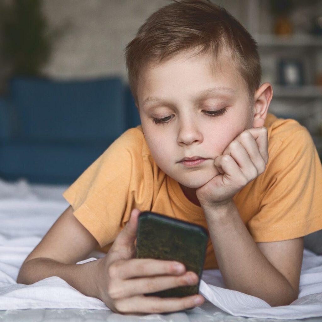 A young boy in a yellow shirt lies on his stomach on a bed, resting his chin on his hand and looking intently at a mobile phone he is holding.